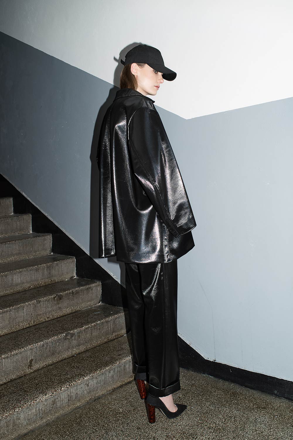 Black lustrous tailoring and a black cap, photographed over the shoulder in a stairwell